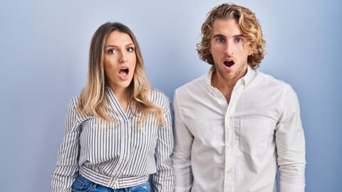 Young couple standing over blue background in shock face, looking skeptical and sarcastic, surprised with open mouth