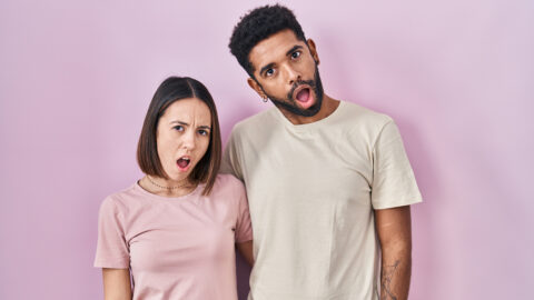 Young hispanic couple together over pink background in shock face, looking skeptical and sarcastic, surprised with open mouth