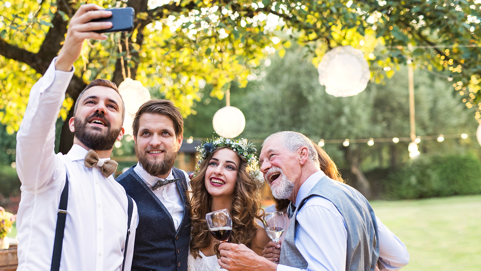 Happy bride, groom and guests with smartphone taking selfie outside at wedding reception.