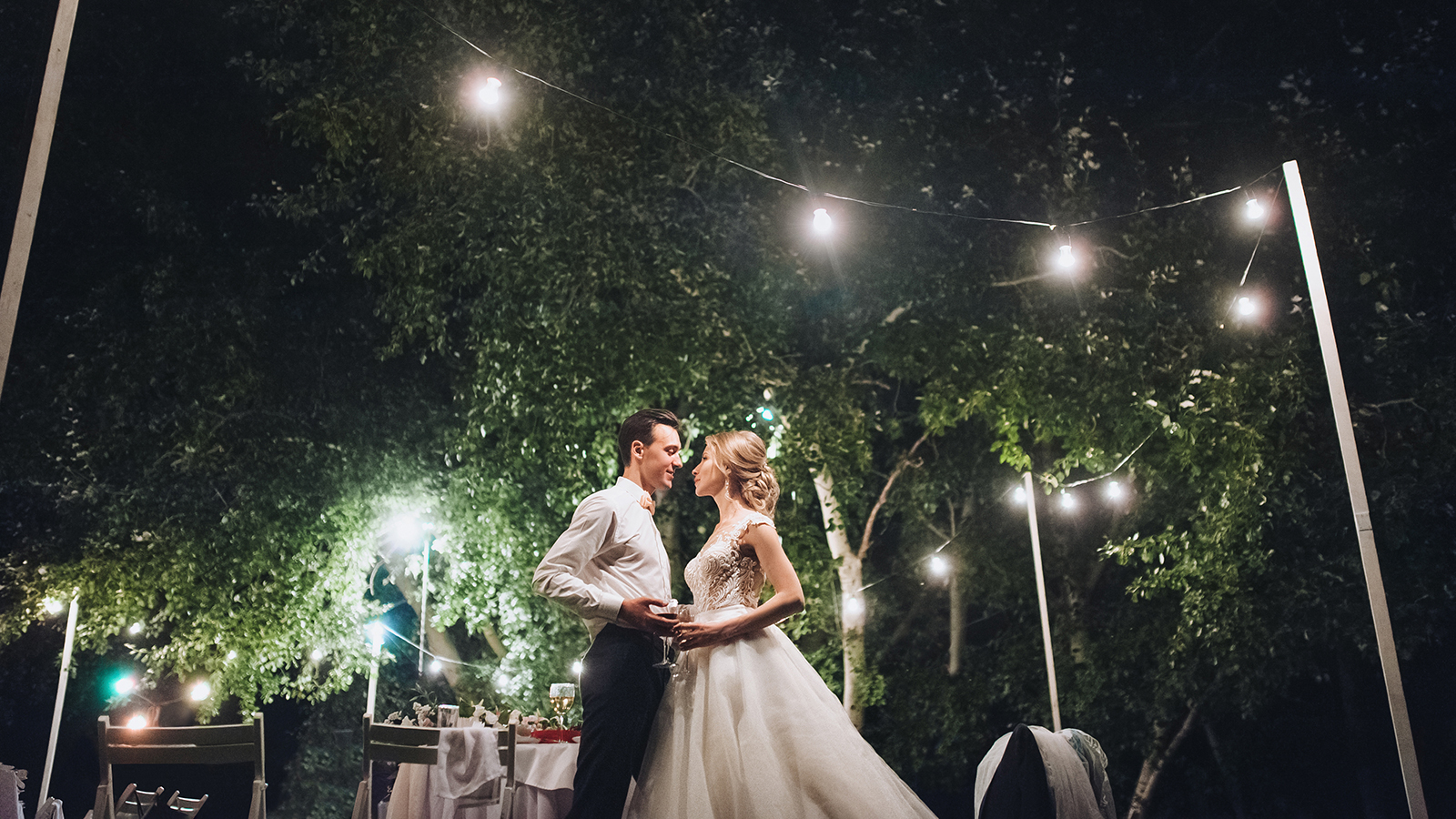 Beautiful newlyweds stand, holding hands, at a wedding party with lamps. Stylish wedding ceremony.