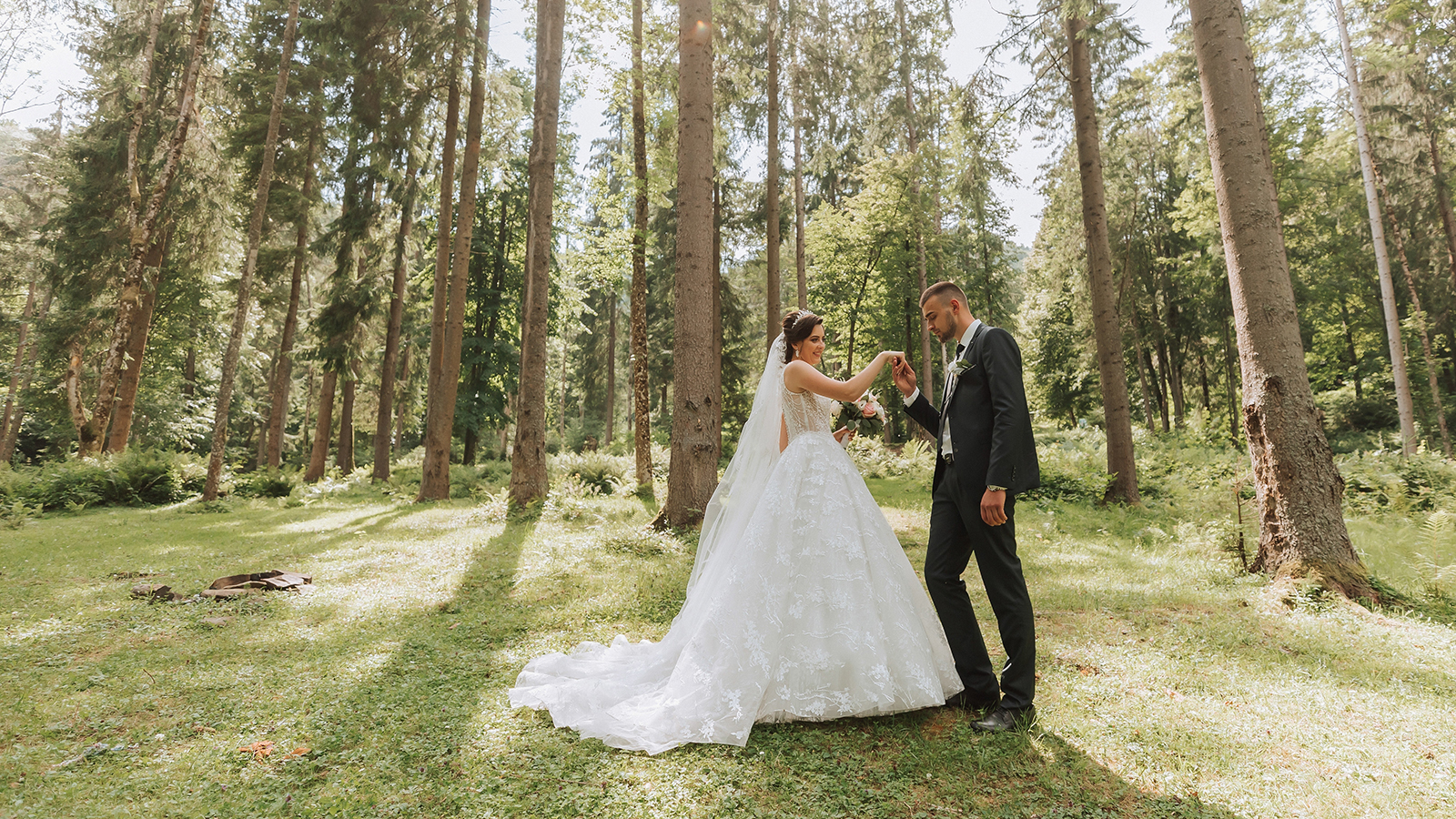Fashionable groom and cute bride in white dress with train and crown on head walking happily in park, garden, forest outdoors. Wedding photography, portrait of smiling newlyweds.