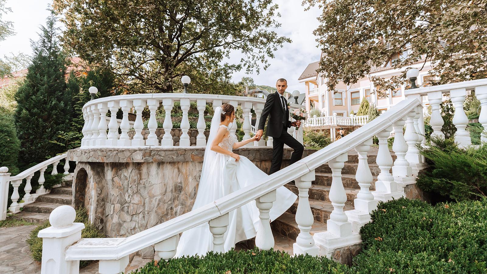 Portrait of a young bride in a white dress and a brunette groom in a suit walking down the steps of an old city park. A beautiful and romantic wedding, a happy couple