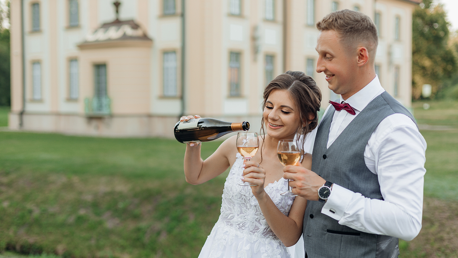 Cheerful married couple of bride in wedding dress and groom in suit pouring and drinking bubbly champagne together, having fun on wedding day in front of new house. Mortgage loan for young family