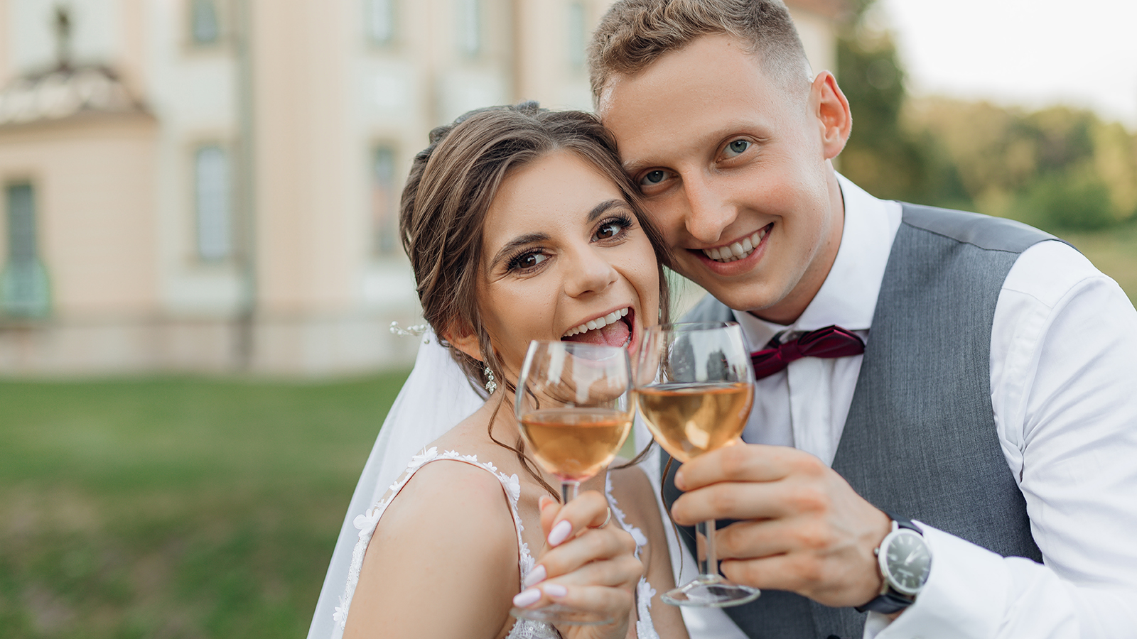 Cheerful, laughing married couple of bride in wedding dress and groom in suit drinking and toasting champagne glasses, having fun on wedding day against new house. Moving new house for young family