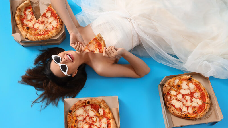 Young woman in white dress with tasty pizza lying on blue background, top view