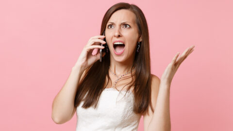 Angry Irritated bride woman in white wedding dress spreading hands, talking on mobile phone, screaming and swearing isolated on pastel pink background. Wedding to do list. Organization of celebration