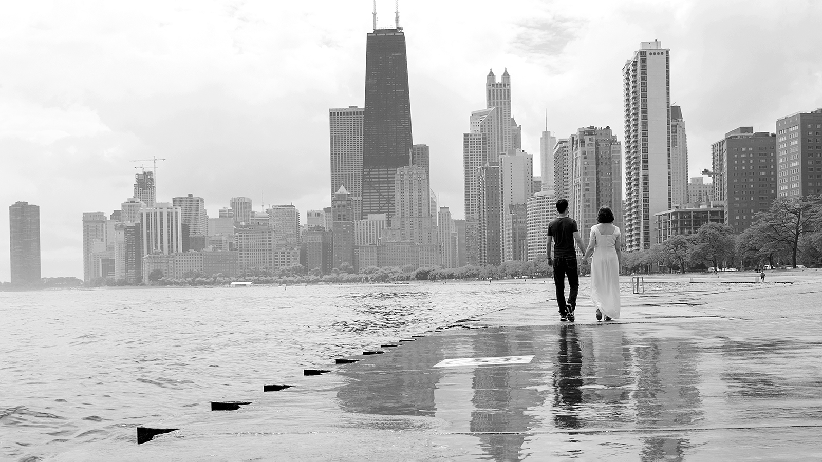 Bride and groom holding hands while walking toward Chicago skyline next to the Lake Michigan in black and white Reflections of the skyscrapers