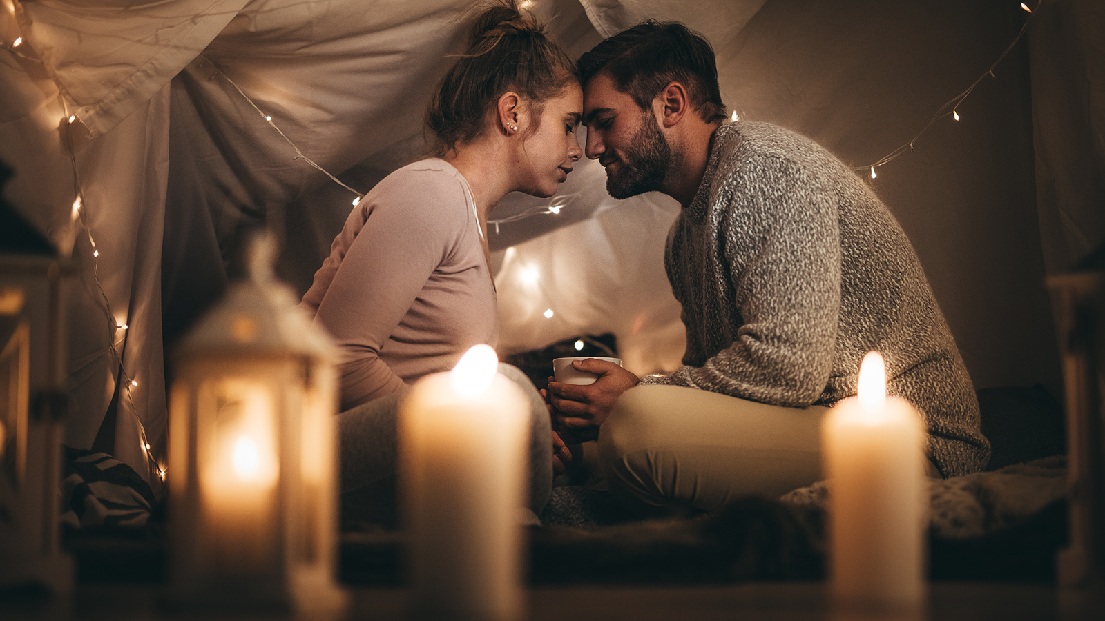 Romantic couple sitting on bed facing each other touching their foreheads and with eyes closed. Man sitting on bed with his wife holding a coffee cup in a room decorated with candles and tiny serial bulbs.