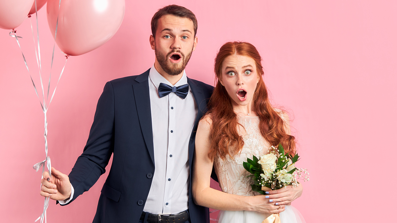 wedding couple posing together isolated over pink background. man in tuxedo holding pink air ballons, woman in white wedding dress bouquet. Woman with auburn hair, couple with opened mouth