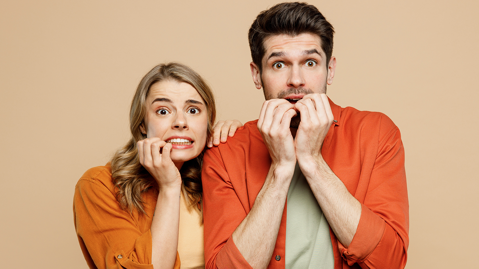 Young shocked scared fearful couple two friends family man woman wear casual clothes looking camera biting nails fingers together isolated on pastel plain light beige color background studio portrait