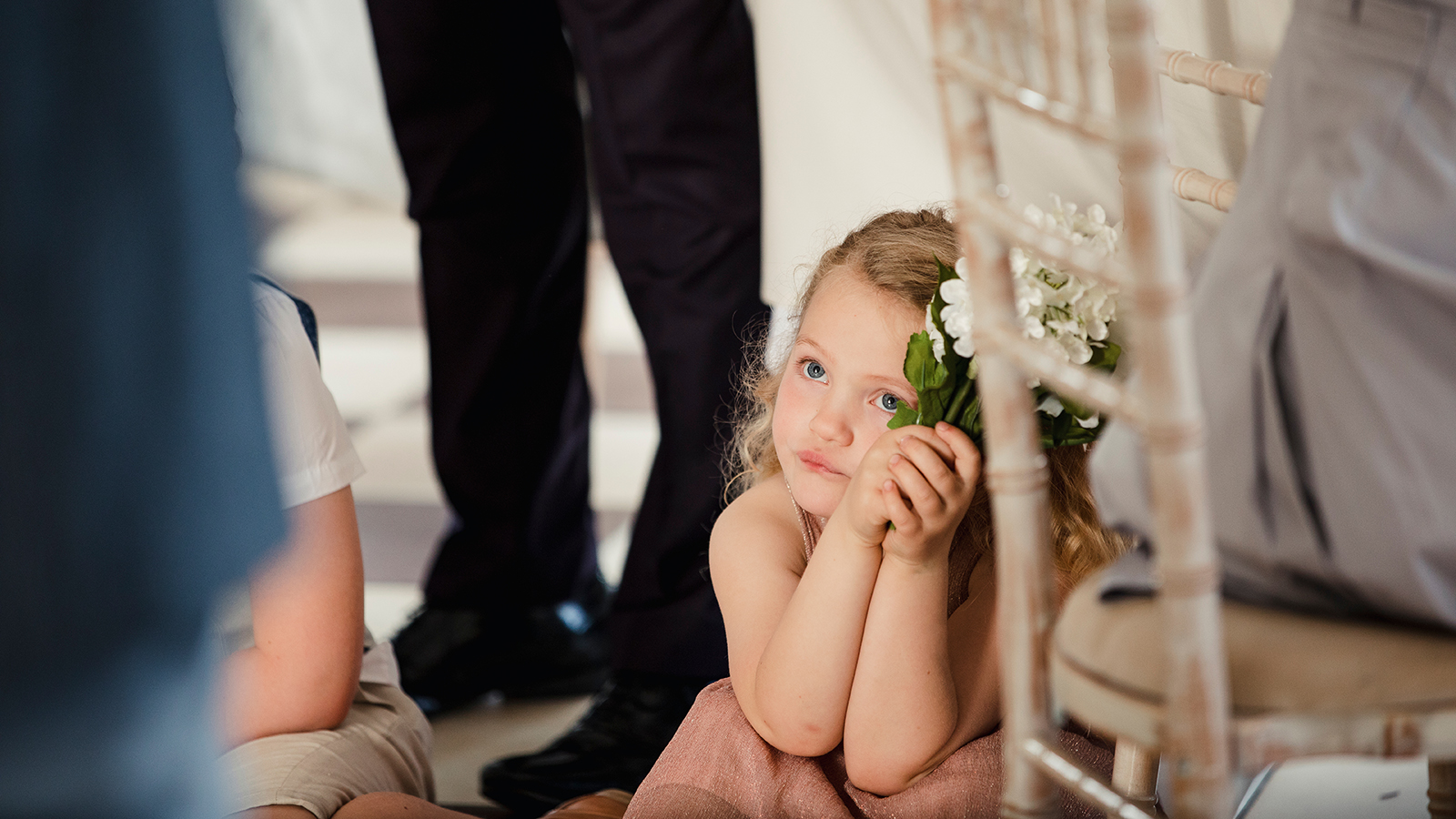 Little girl is sitting on the dancefloor by a table at a wedding. She is watching the bride and groom share their first dance.
