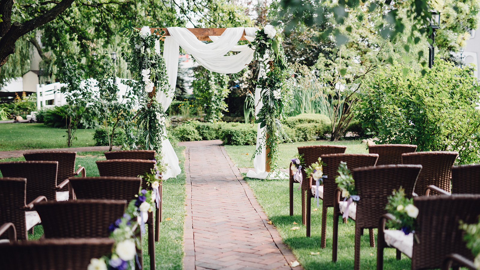 Wooden wedding arch decorated by white cloth and flowers with greenery standing in the center of wedding ceremony. Brown chairs on side.