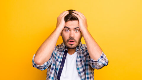 Portrait of frightened brunette man in a plaid shirt holds his hands behind his head and looks into the camera isolated on shine yellow background with copy space for text