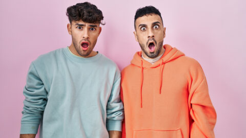Young hispanic gay couple standing over pink background in shock face, looking skeptical and sarcastic, surprised with open mouth