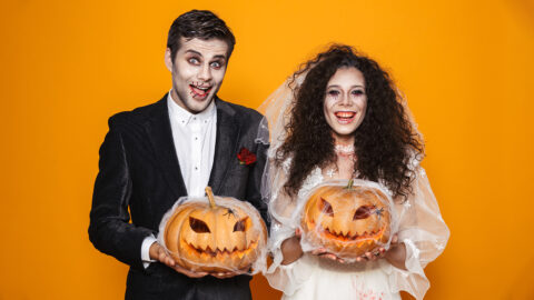 Photo of horrific zombie couple bridegroom and bride wearing wedding outfit and halloween makeup holding carved pumpkin isolated over yellow background