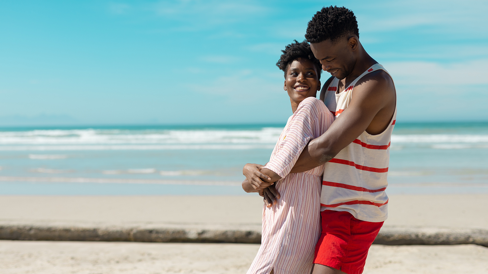 Side view of african american young man embracing smiling girlfriend at beach against blue sky. nature, summer, unaltered, beach, love, togetherness, lifestyle, enjoyment and holiday concept.
