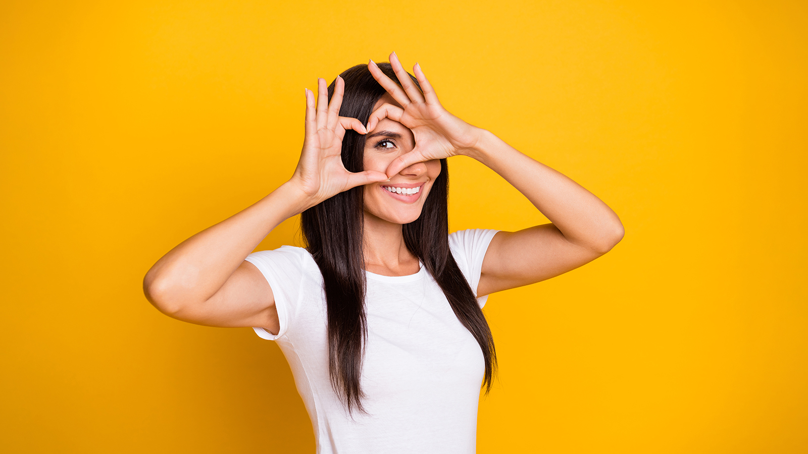 Photo portrait of brunette girl showing heart shape love with fingers near face looking smiling isolated on vibrant yellow color background.