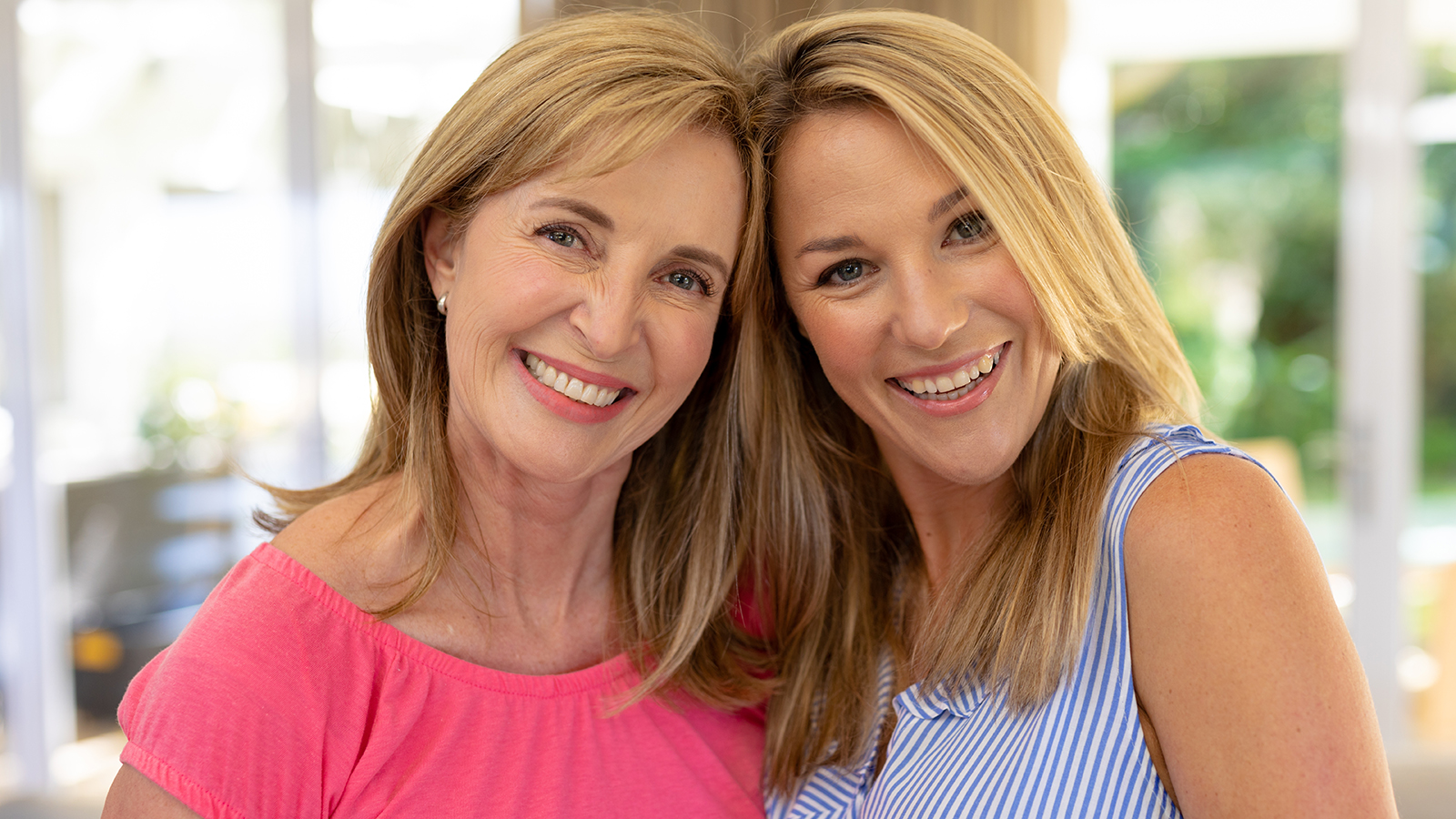 Portrait close up of a Caucasian woman and her adult daughter at home, standing in the living room embracing, touching heads together and looking to camera smiling