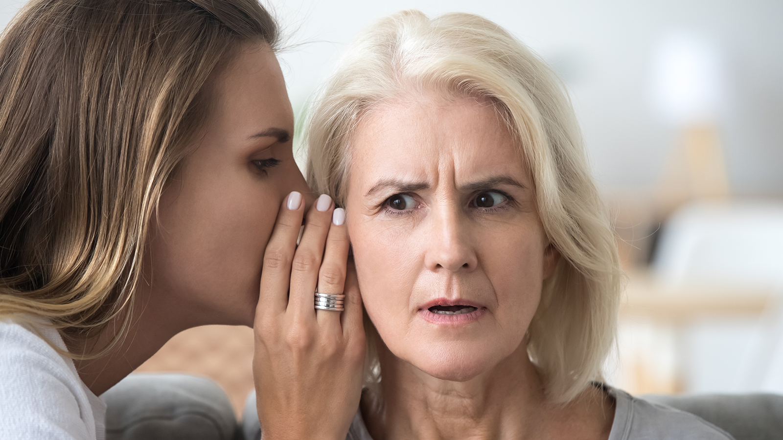 Young woman whisper in aged mother ear sharing secret or hidden information, surprised elderly mom hearing shocking news from adult daughter, parent and kid being best friends gossiping at home