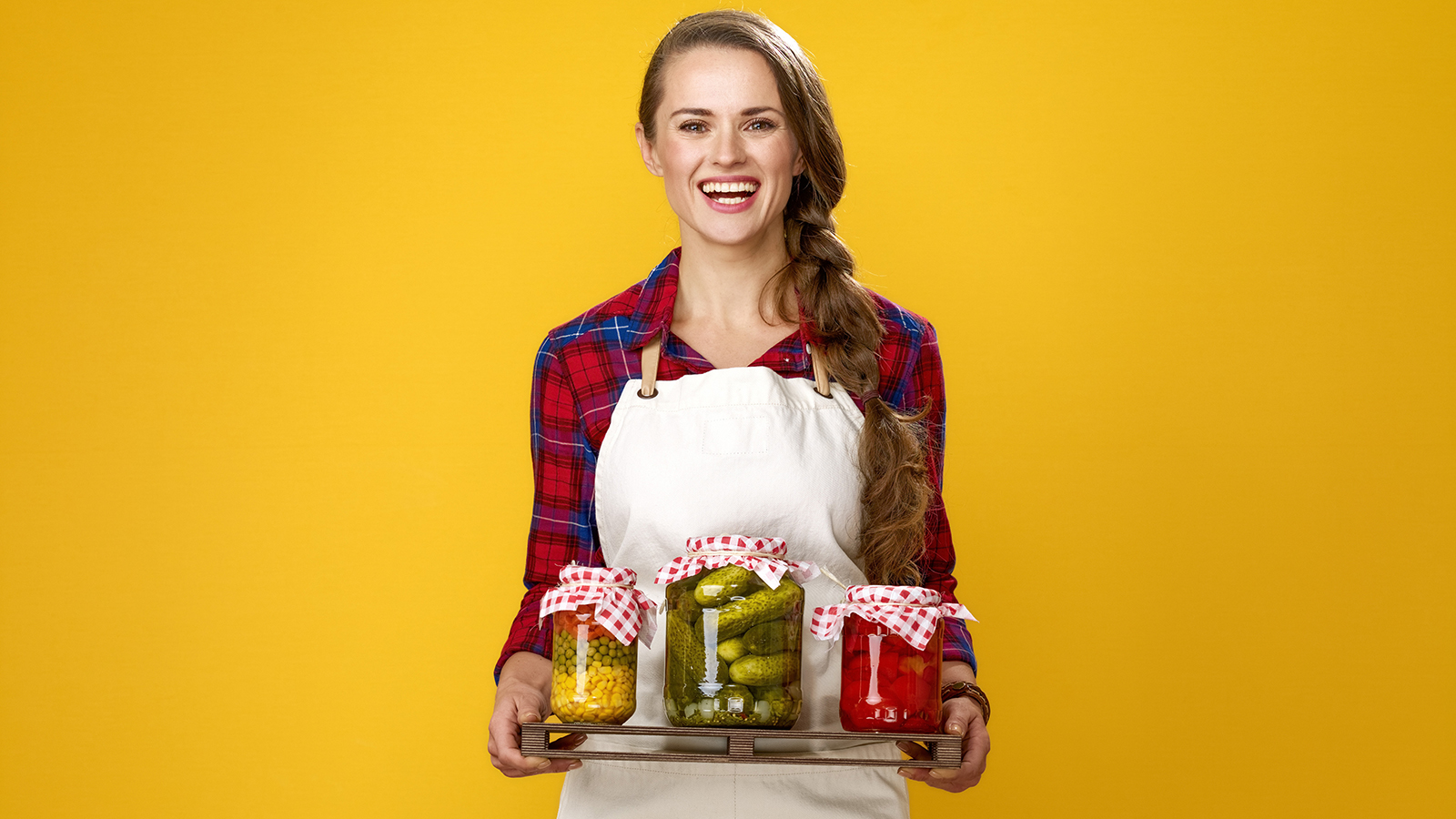 Healthy food to your table. Portrait of happy young woman cook wearing apron isolated on yellow showing a jars of marinated vegetables
