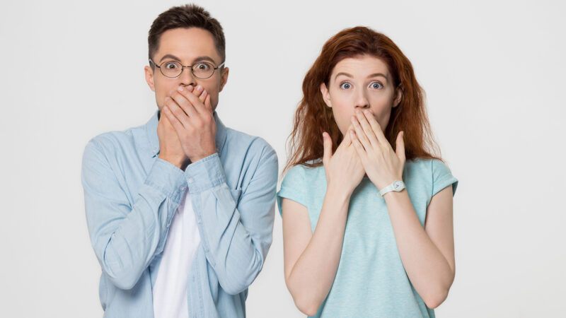 Shocked scared amazed couple covering mouth with hands feel horrified stunned looking at camera, surprised mute man and woman astonished about unexpected news isolated on white grey studio background