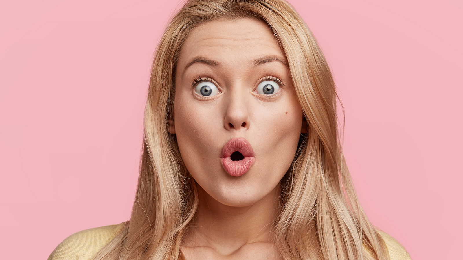 Puzzled shocked female looks with bugged eyes at camera, being surprised to see big sales, dressed casually, isolated over pink studio background. Emotional frightened woman indoor afraid of something