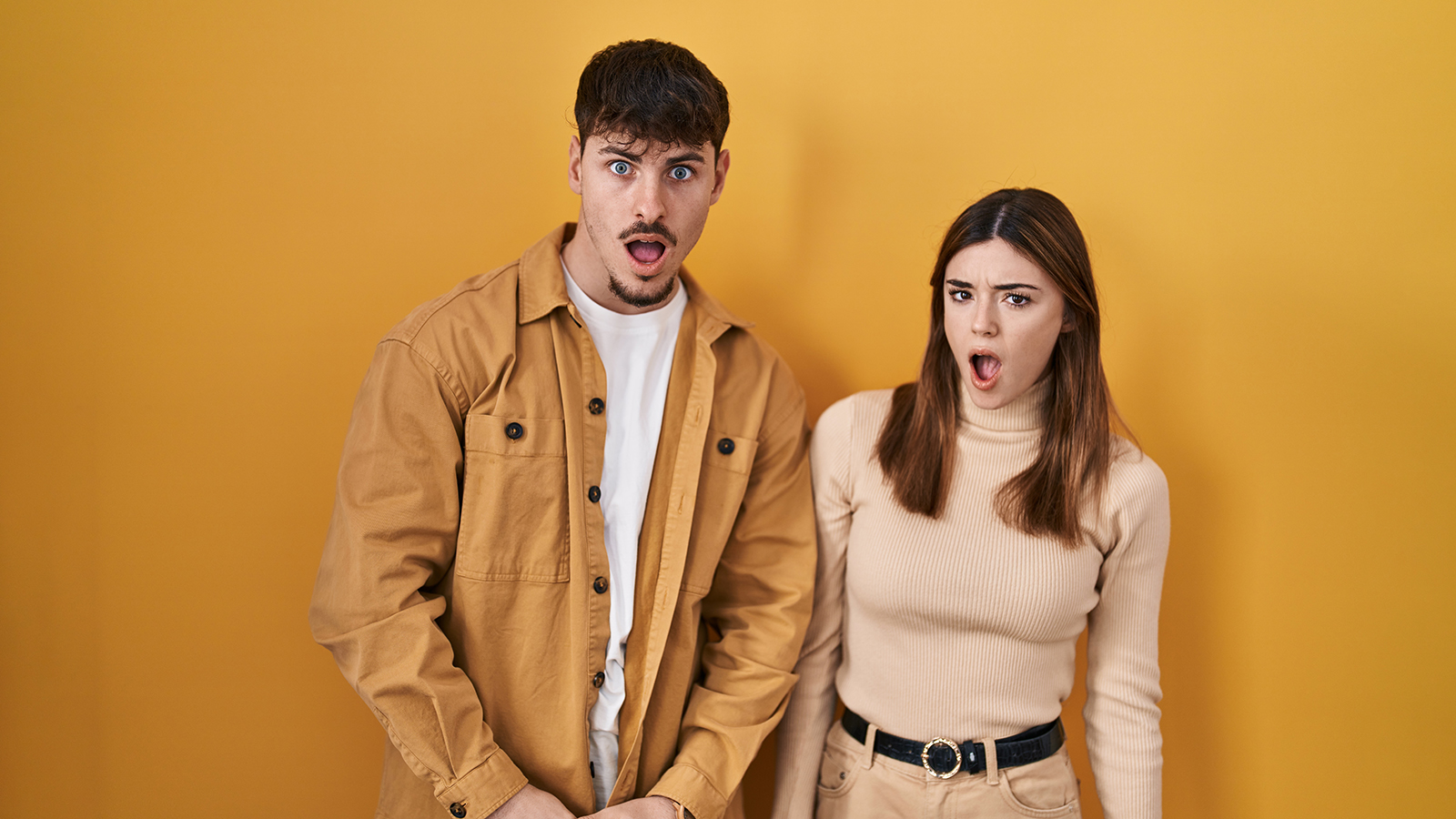 Young hispanic couple standing over yellow background in shock face, looking skeptical and sarcastic, surprised with open mouth