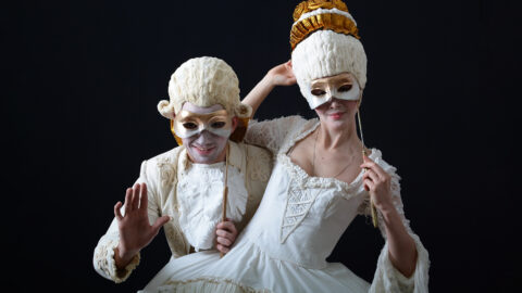 a man and a woman in period costume and wigs and holding a theatrical mask