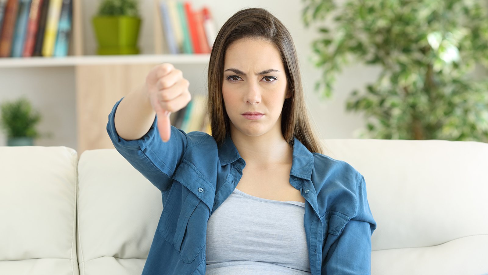 Angry woman looking at camera with thumbs down sitting on a couch in the living room at home