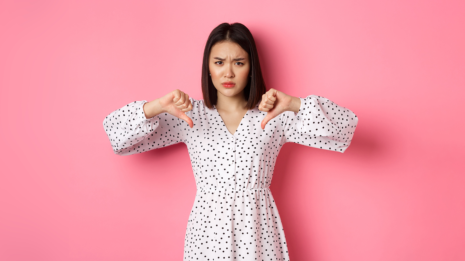 Disappointed asian woman showing thumbs-down, disapprove and dislike something, showing negative judgement, standing over pink background.