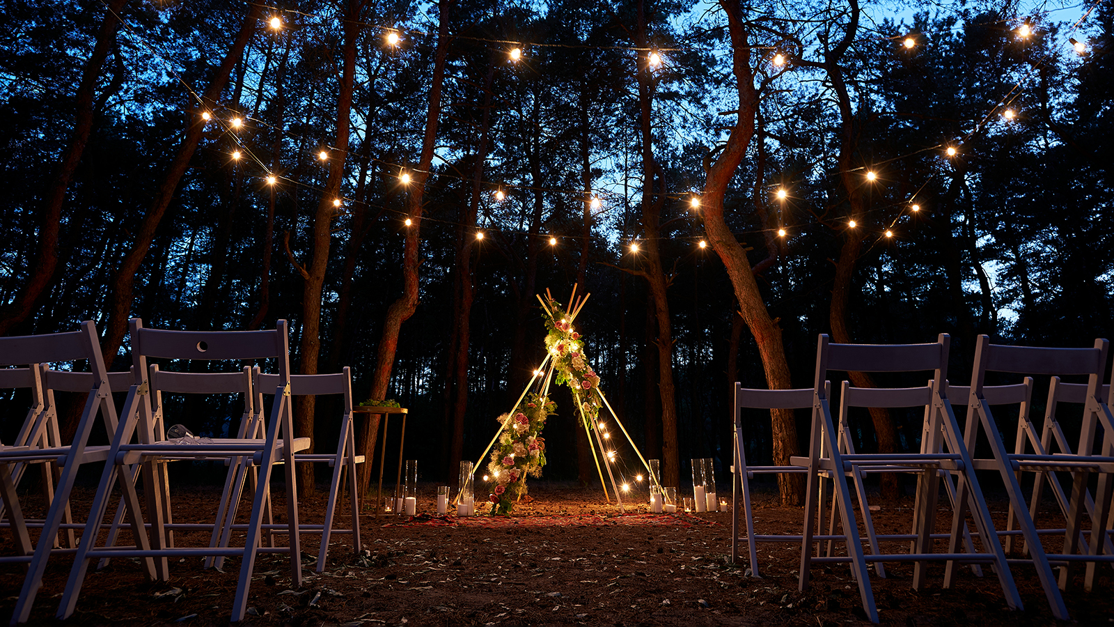 Festive string lights illumination on boho tipi arch decor on outdoor wedding ceremony venue in pine forest at night. Vintage string lights bulb garlands shining above chairs at a summer rural wedding