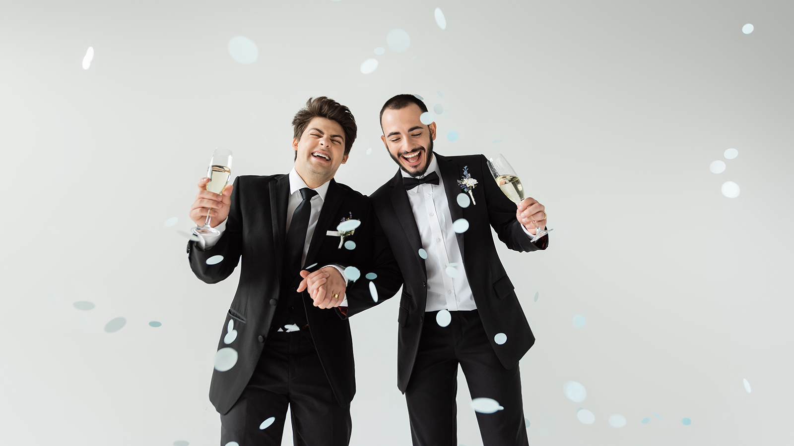 Excited homosexual grooms in elegant formal wear holding hands and glasses of champagne while standing under falling confetti during wedding on grey background