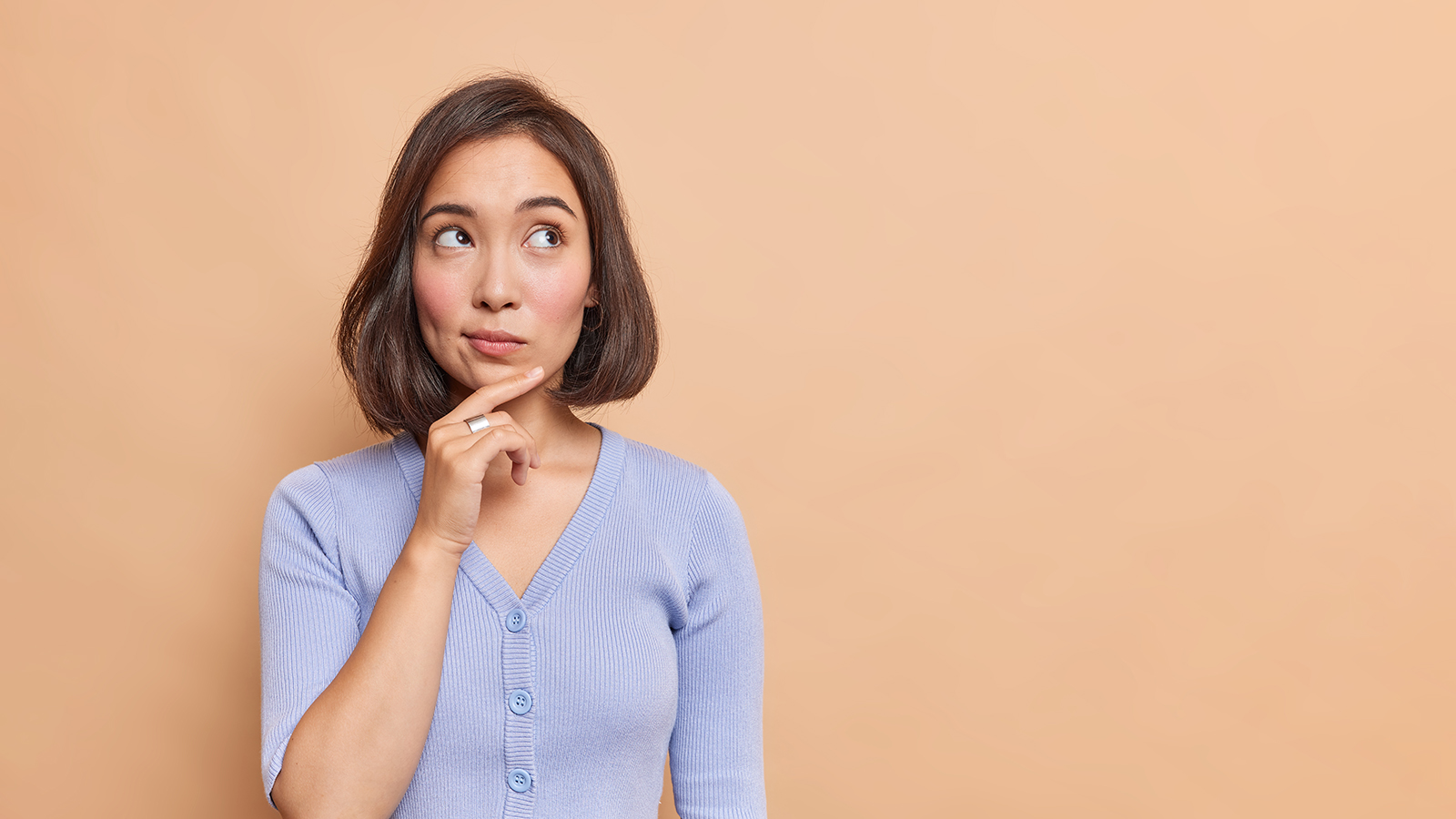 Thoughtful Asian woman keeps hand on chin looks pensively above dressed in casual blue jumper poses against brown background blank copy space for your advertising content thinks about future