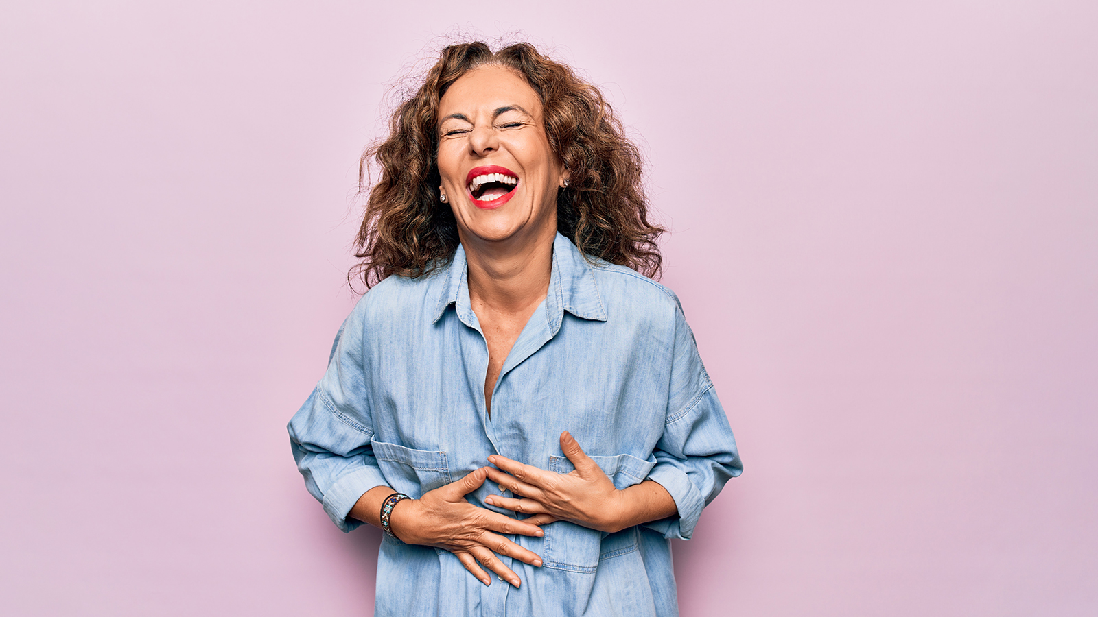 Middle age beautiful woman wearing casual denim shirt standing over pink background smiling and laughing hard out loud because funny crazy joke with hands on body.