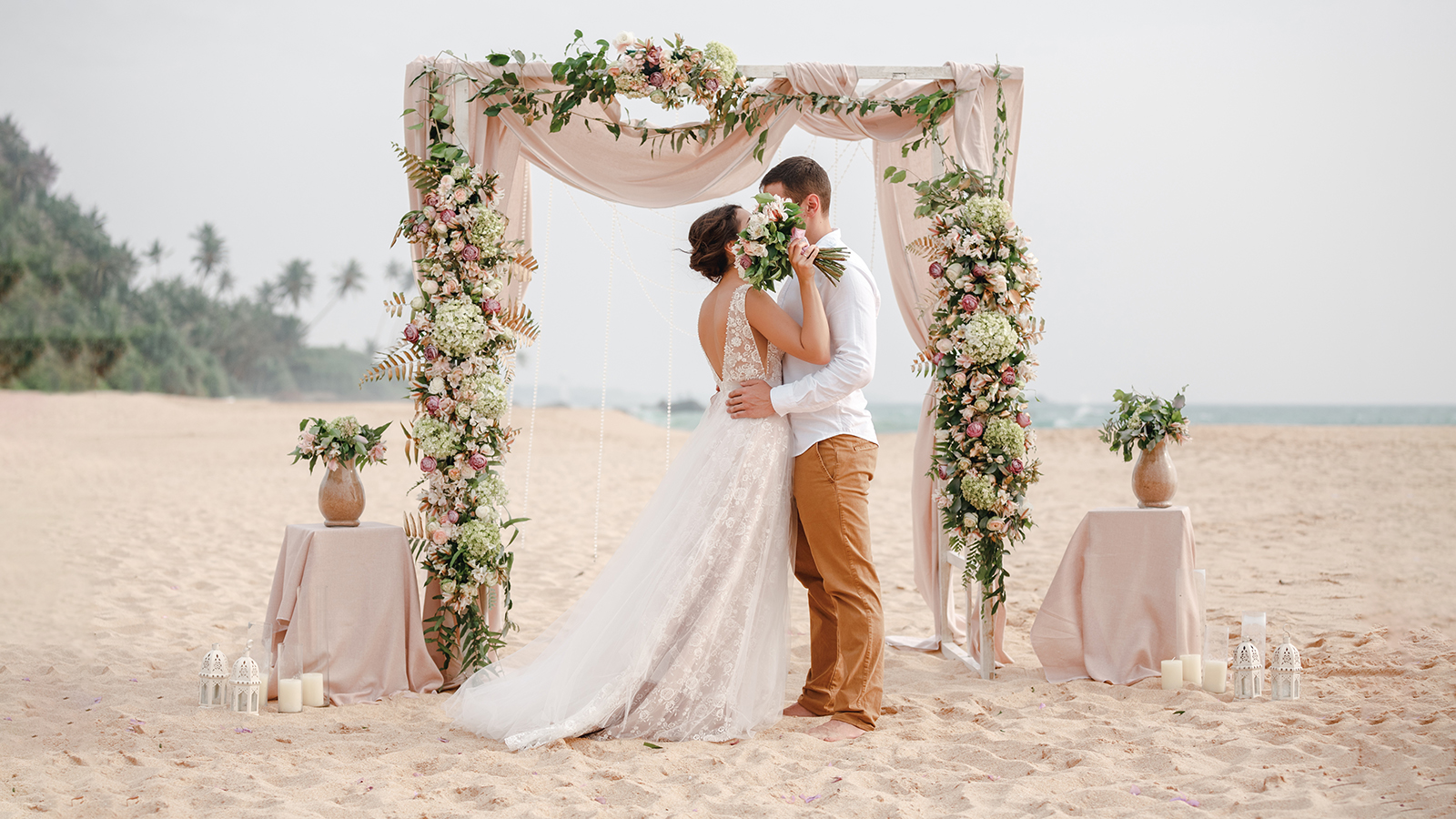 Bride and groom enjoying beach wedding in tropics, wedding arch, ocean background. Wedding ceremony on a tropical beach. Happy groom and beautiful bride kissing under the arch decorated with flowers