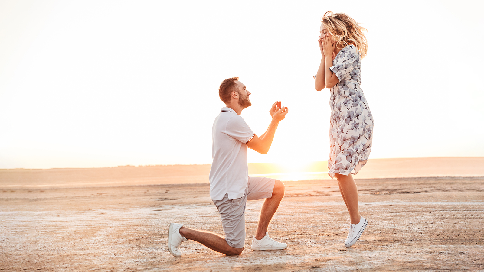 Photo of happy man making proposal to his excited woman with ring in gift box while walking on sunny beach