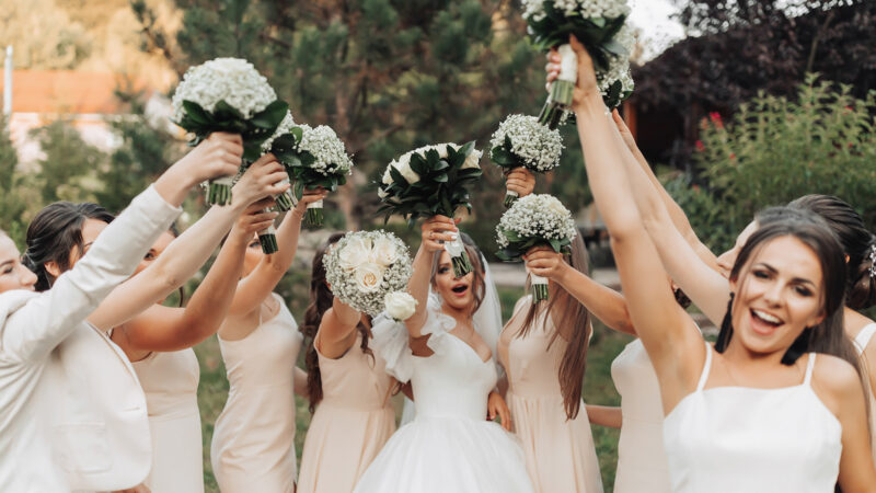 Wedding photo in nature. A brunette bride in a white long dress and her friends in nude dresses are standing against the background of trees, smiling, holding up their gypsophila bouquets. Young women