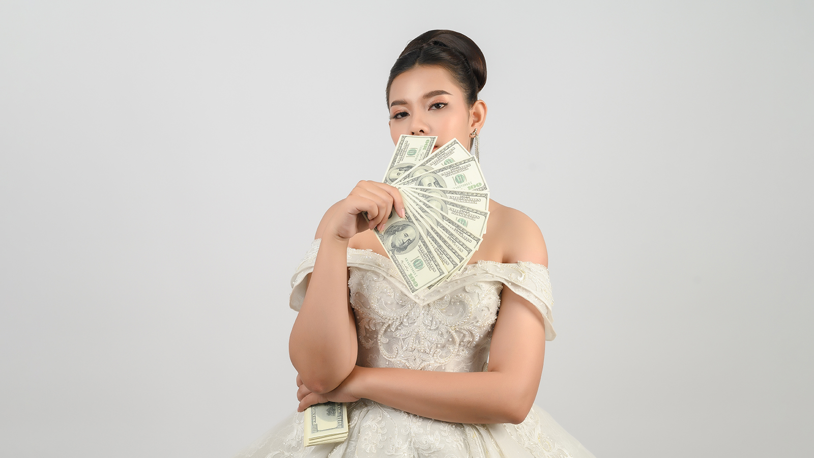 Portrait Young asian beautiful bride in white wedding dress holding dollar bills fan in hand closed her mouth on white background, looking camare