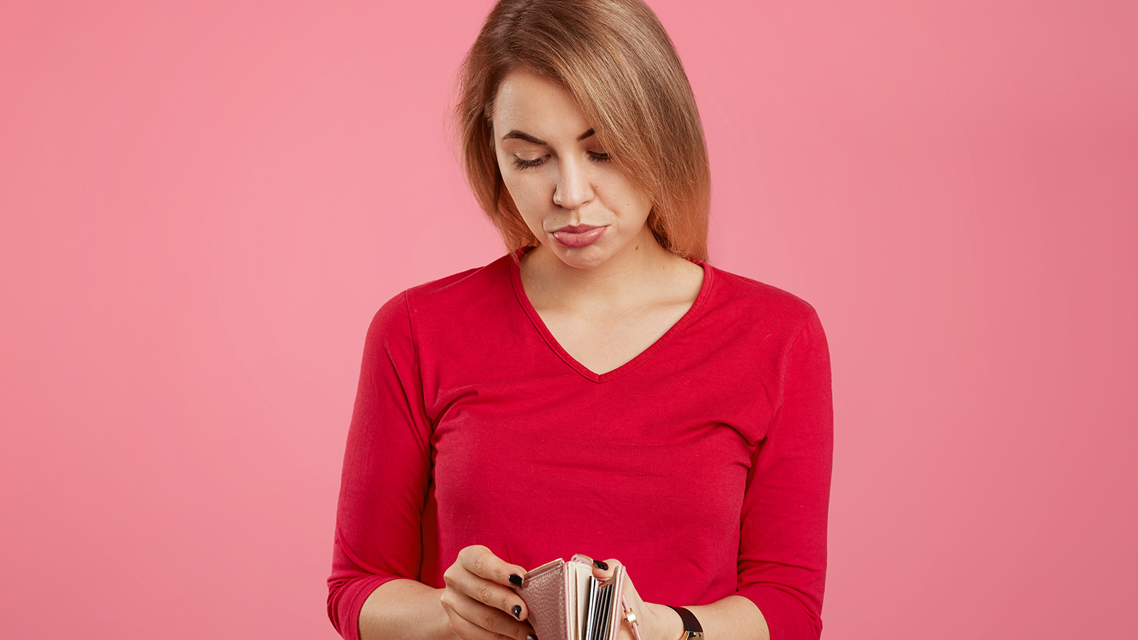 Bankruptancy and finance concept. Displeased attractive female looks with unhappy expression in purse, has no money and many debts, curves lower lip, isolated over pink background. Empty wallet