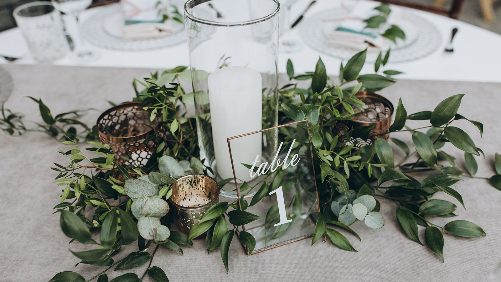on table with gray tablecloth is a candle in candlestick, plate with number and composition of greenery