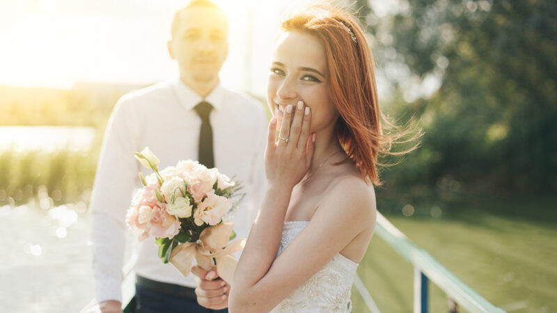 redhead bride in white dress holding flower bouqet and groom in white shirt and tie are standing on the bridge in the wedding summer day. Woman is pleasantly surprised by the gift