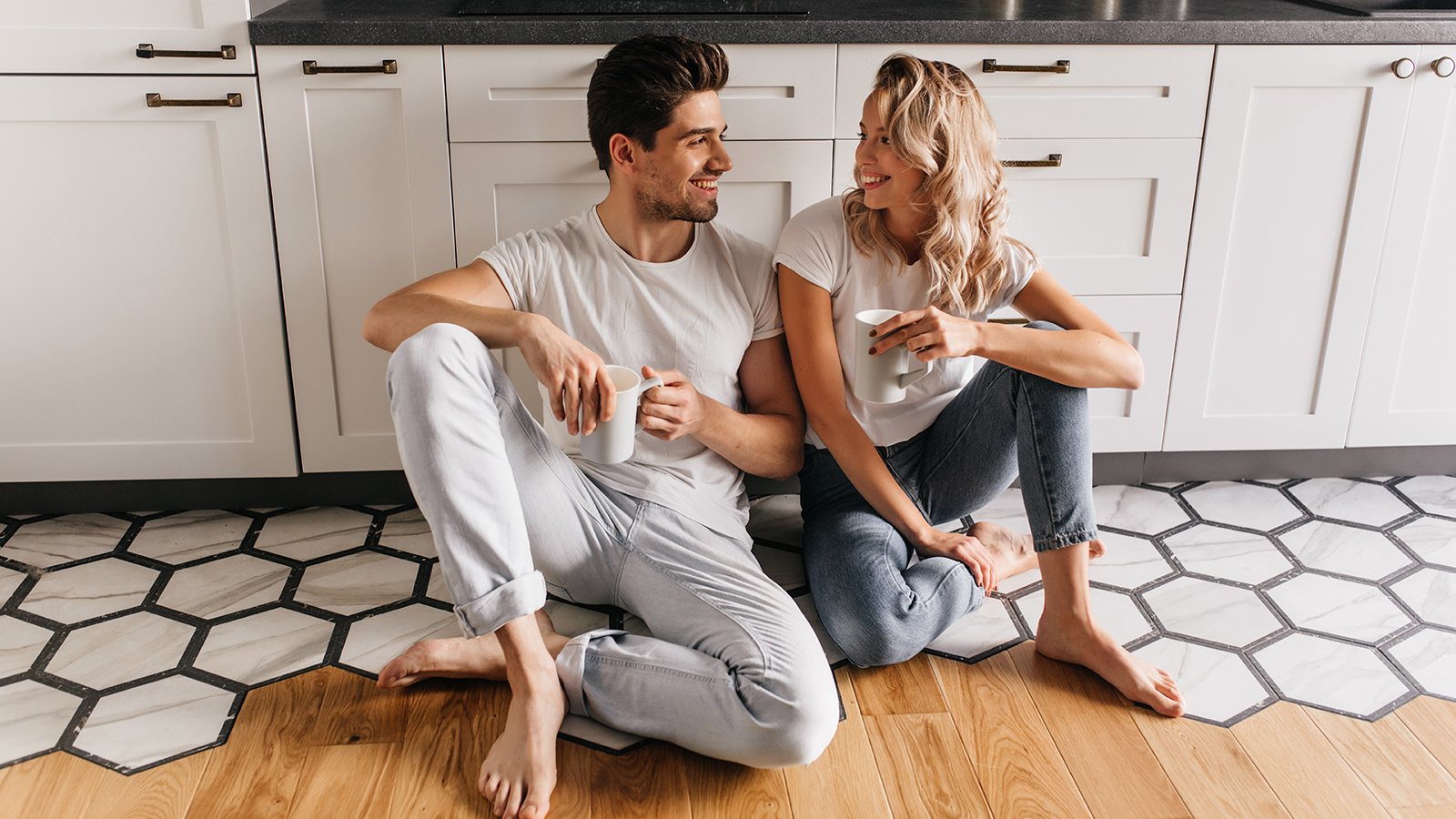 Lovely girl in jeans sitting on the floor and talking with boyfriend. Young couple enjoying coffee in kitchen.