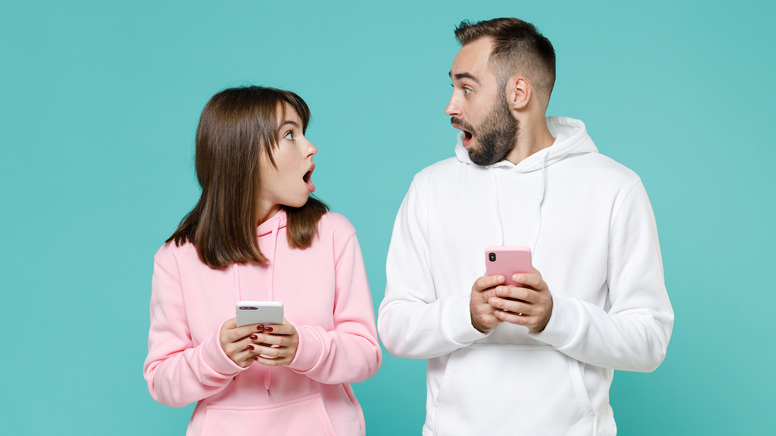 Shocked young couple two friends man woman 20s wearing white pink casual hoodie using mobile cell phone typing sms message looking at each other isolated on blue turquoise background studio portrait