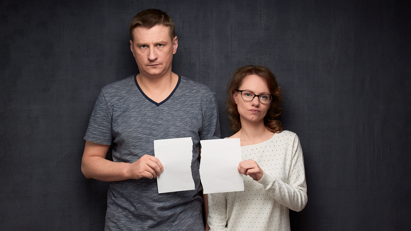 Studio half-length portrait of dissatisfied caucasian couple dressed casually, holding parts of torn white paper sheet in hands, looking with displeasure at camera, standing over gray background