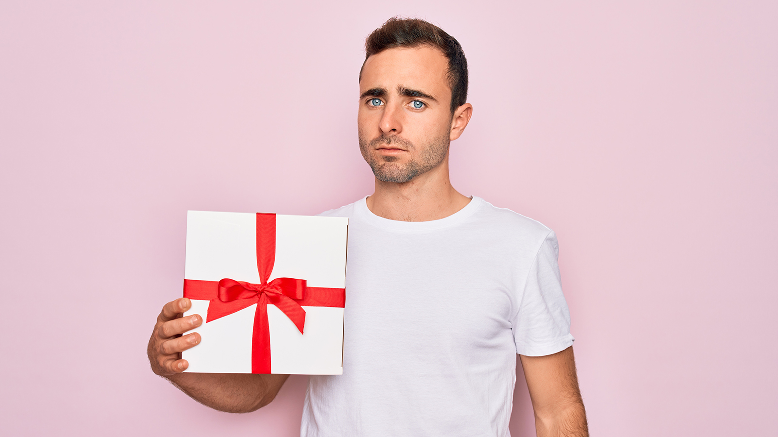 Young handsome man with blue eyes holding birthday present over isolated pink background with a confident expression on smart face thinking serious