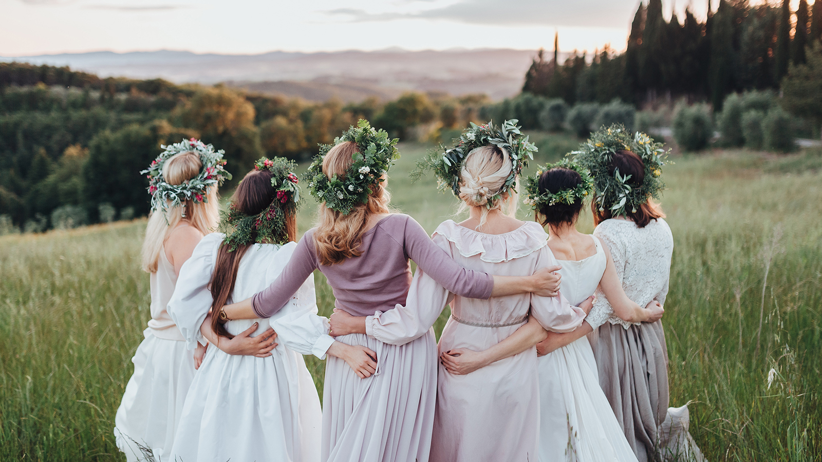 Girls in dresses and wreaths of flowers and greenery are in the green field at sunset