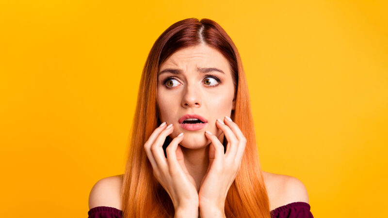 Oh no! Human emotions concept. Close up portrait of confused beautiful woman with open mouth big eyes isolated on yellow background. Young scared girl touch one's face with hands and look aside