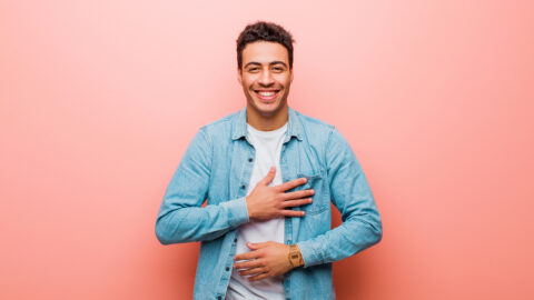 young arabian man laughing out loud at some hilarious joke, feeling happy and cheerful, having fun against pink wall
