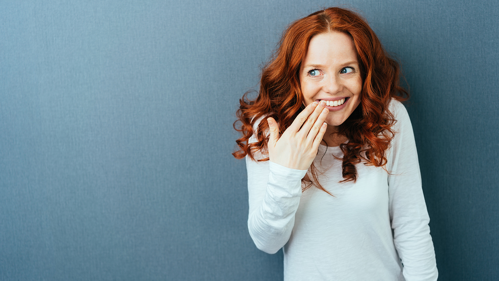 Cute young woman with gorgeous long curly red hair standing laughing behind her hand over a dark studio background with copy space