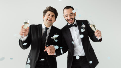 Excited gay grooms in elegant formal wear holding hands and glasses of champagne while standing under falling confetti during wedding on grey background
